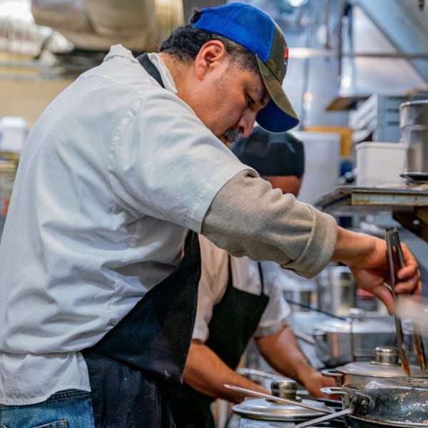 A chef in a blue cap and white shirt cooking with tongs in a busy Mexican restaurant kitchen.