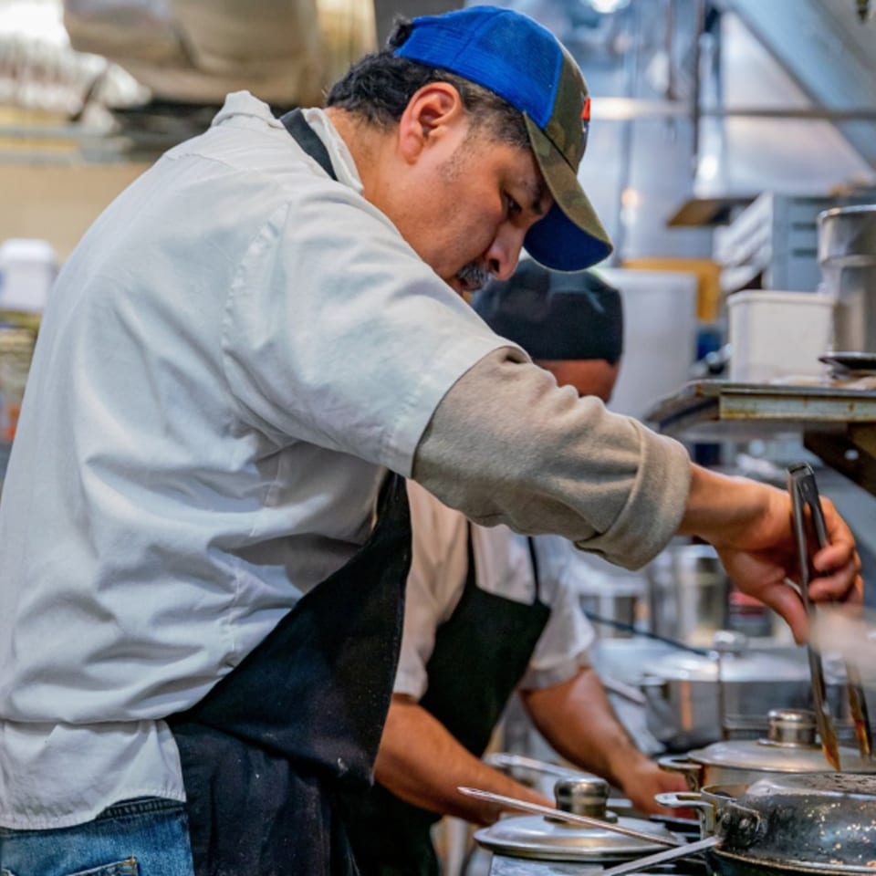 A chef in a blue cap and white shirt cooking with tongs in a busy Mexican restaurant kitchen.