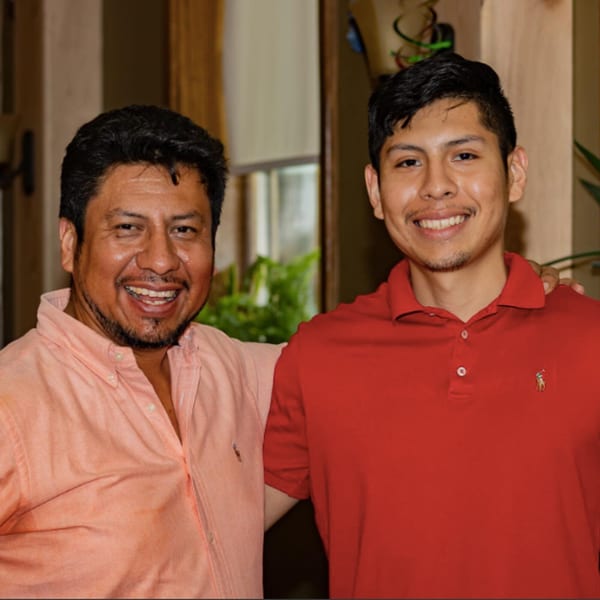 Two smiling men, one in a peach shirt and one in a red polo, stand together.