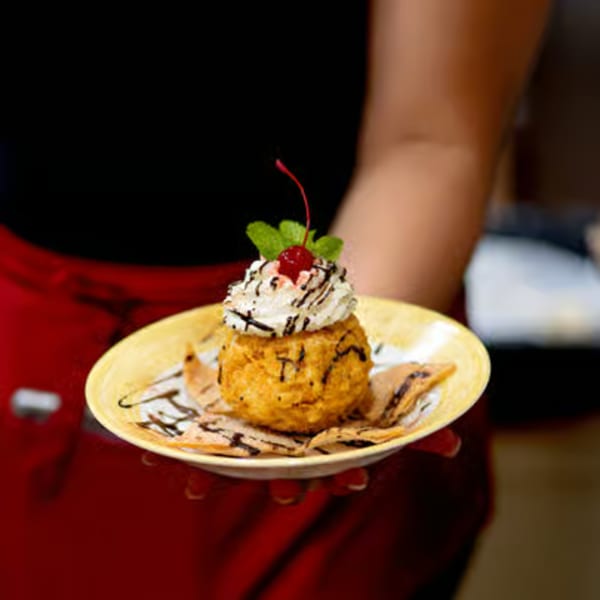 A fried ice cream dessert with whipped cream, chocolate drizzle, cherry, and mint, held by a server.