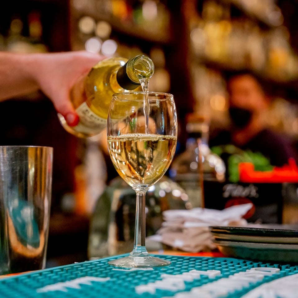 A hand pours white wine from a bottle into a glass on a bar with blurred bottles in the background.