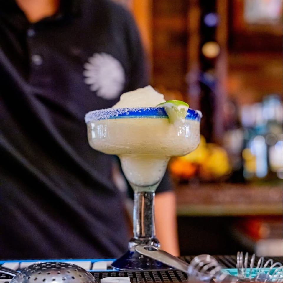 A frozen margarita with a salted blue rim and lime wedge, on a bar with cocktail tools.