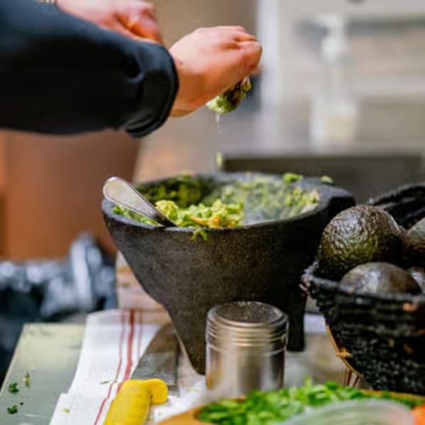 Hands squeezing lime into a molcajete filled with guacamole, alongside fresh avocados.