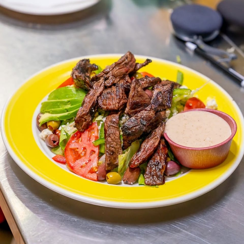 Gaby's Cafe steak salad with avocado, tomatoes, olives, and dressing on a yellow plate.