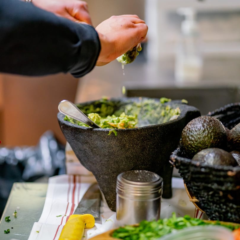 Hands squeezing lime juice into a molcajete filled with guacamole, alongside fresh avocados.
