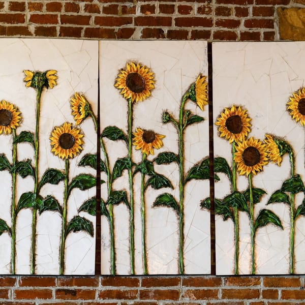 Three panels of sunflower artwork with green stems and leaves on a white background above a brick wall.