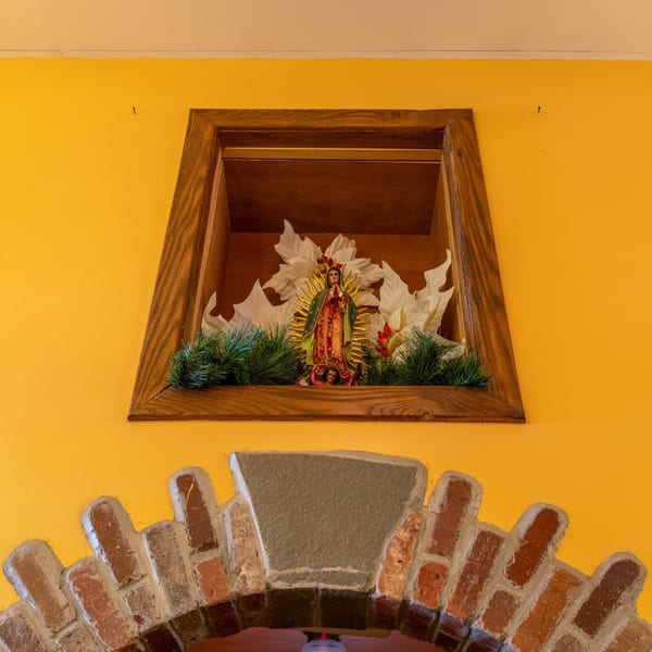 A Virgin Mary statue in a wooden alcove with white flowers and green foliage, above a brick archway.