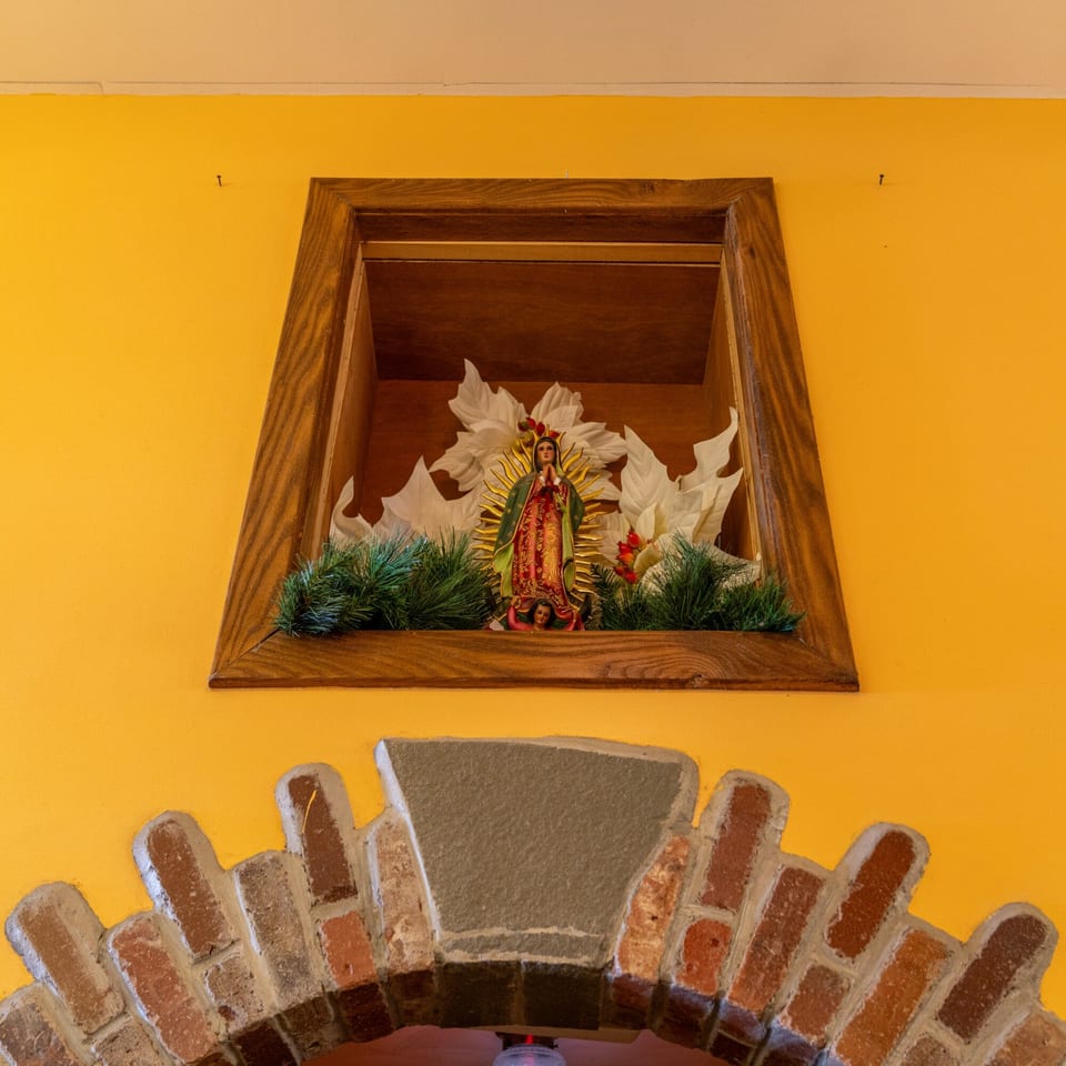 A Virgin Mary statue in a wooden alcove with white flowers and green foliage, above a brick archway.