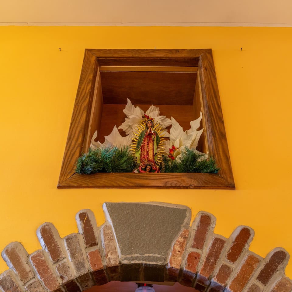 A Virgin Mary statue in a wooden alcove with white flowers and green foliage, above a brick archway.