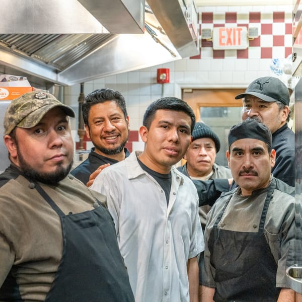 Six Gaby's Cafe kitchen staff members, wearing aprons and hats, pose together in the kitchen.