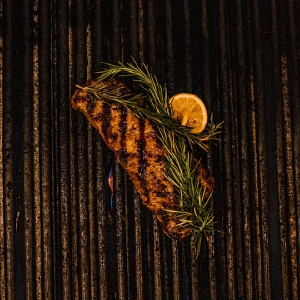 Grilled steak with rosemary and a lemon slice on a dark grill, with a blue flame visible.