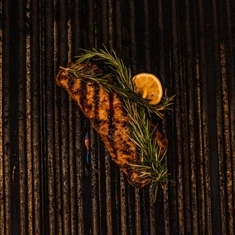 Grilled steak with rosemary and a lemon slice on a dark grill, with a blue flame visible.